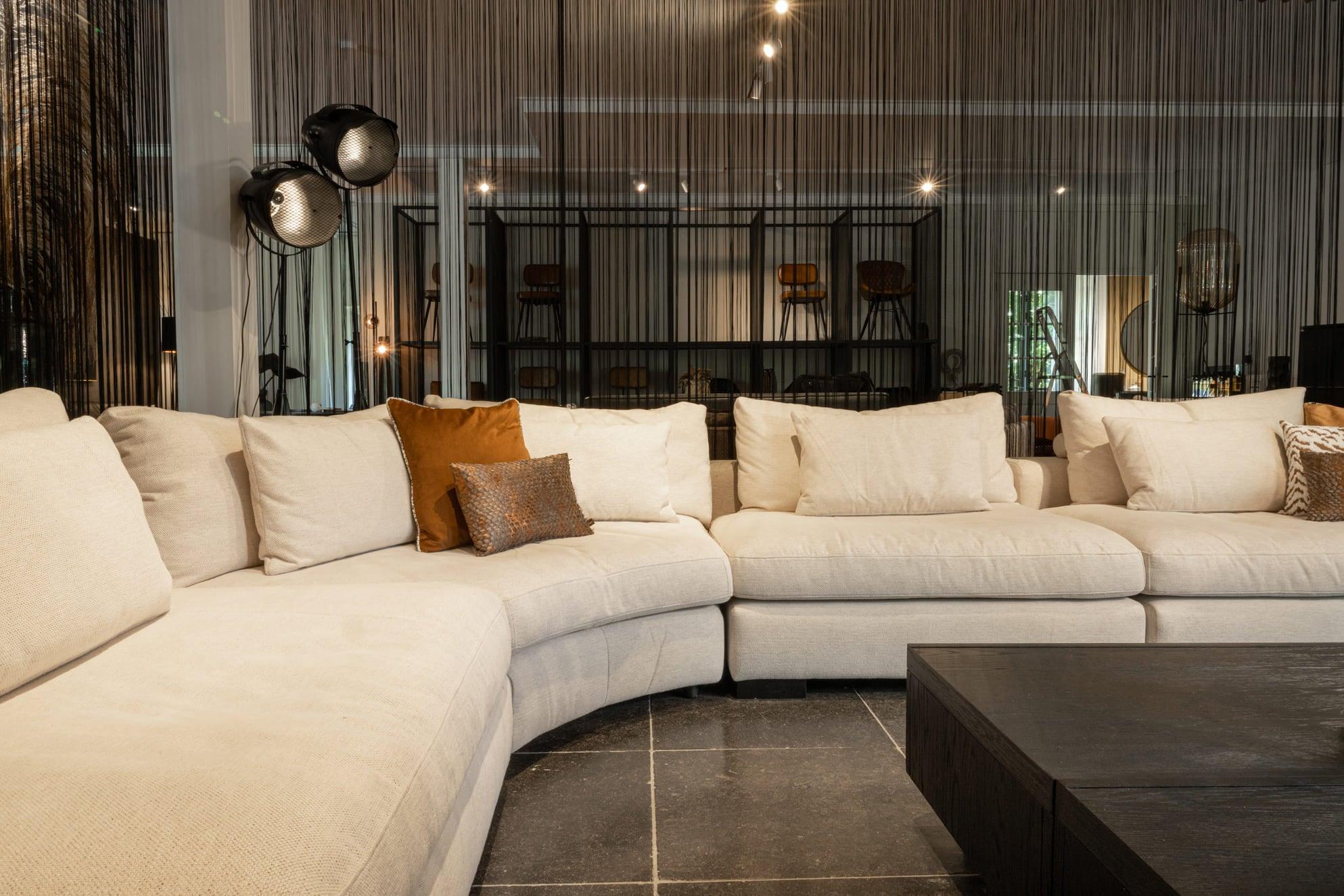 "Gestoffeerde beige hoekbank met sierkussens in stijlvol ingerichte woonkamer met industriële boekenkast, design vloerlamp en moderne decoratie."