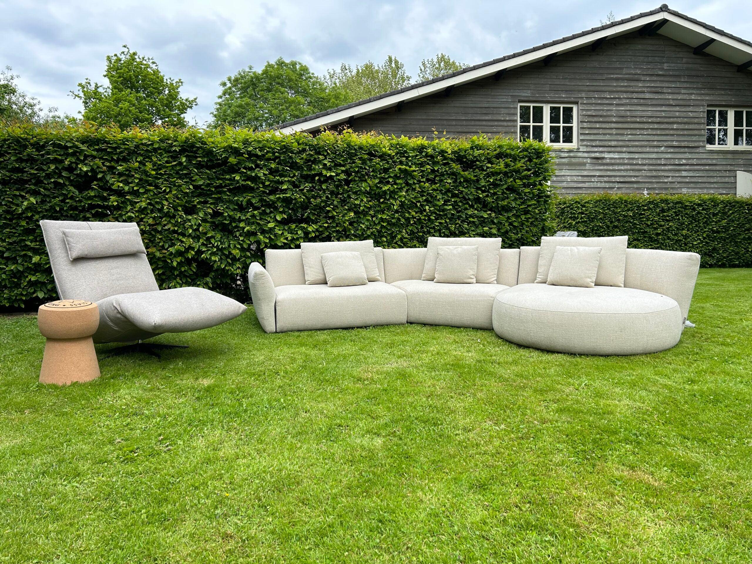 "Moderne buitenlounge set met een chaise longue en kussens op een netjes onderhouden gazon tegenover een donker houten tuinschuur omgeven door groene heggen."