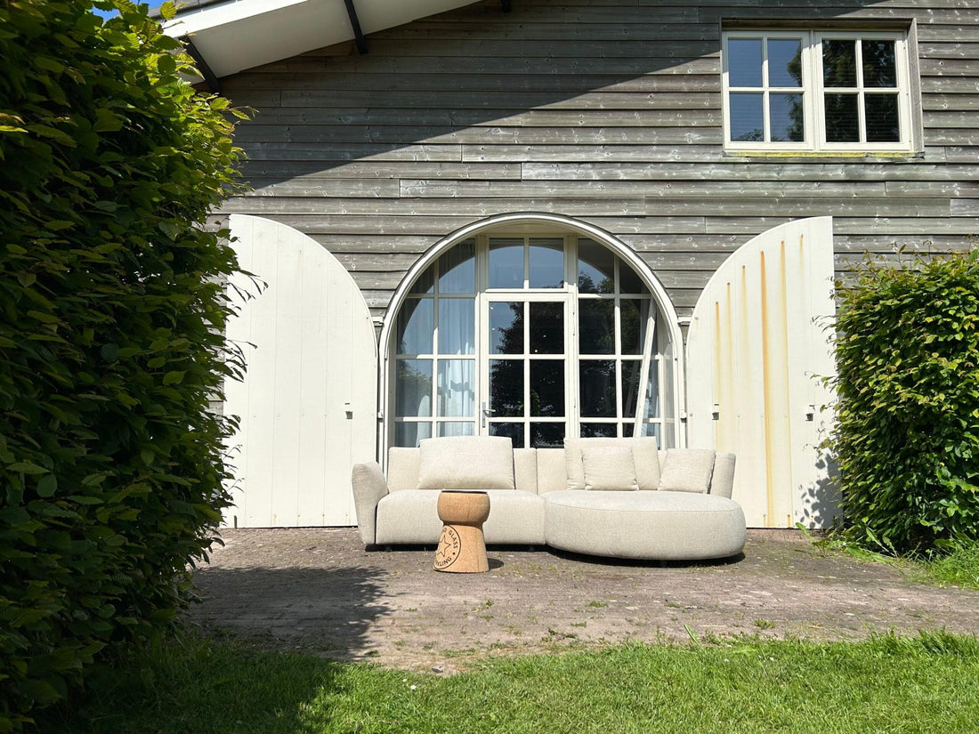 "Beige modulaire hoekbank voor landhuis met grote boogramen en openslaande deuren, omgeven door groene beplanting onder zonnige hemel."