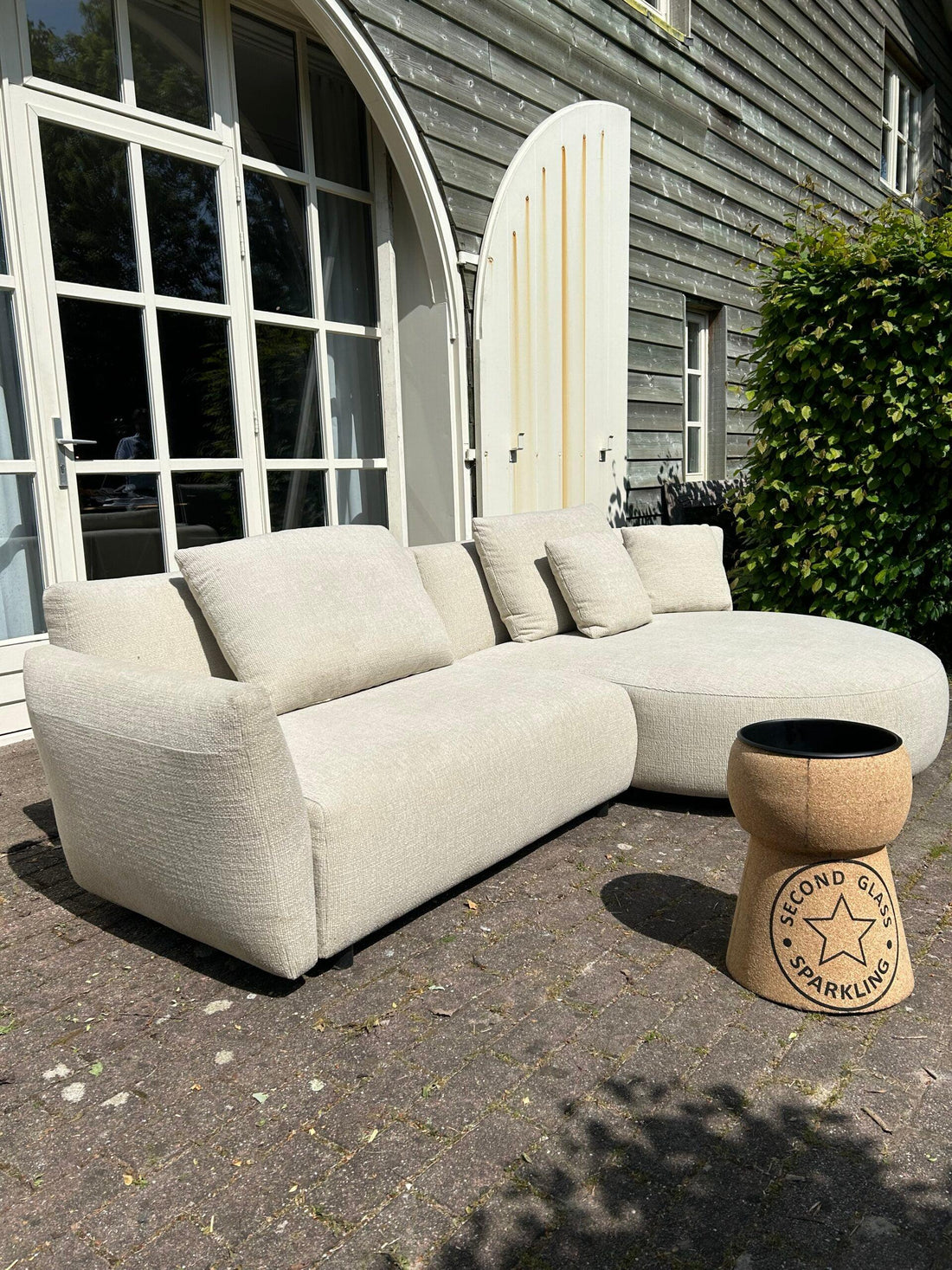 "Beige comfortabele loungebank voor buiten tegen achtergrond van houten woning met grote boogvormige witte deuren, vergezeld van een unieke bijzettafel in de vorm van een kurk, op een geplaveide terras met groene beplanting."