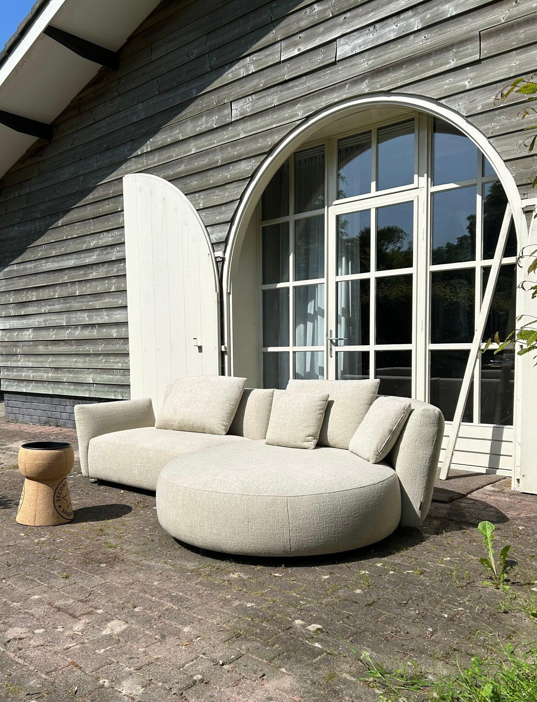 "Ronde designer loungebank voor buiten in beige stof, geplaatst voor een houten huis met grote boogvormige ramen en openstaande witte deur, naast een decoratieve keramische bijzettafel met patronen."