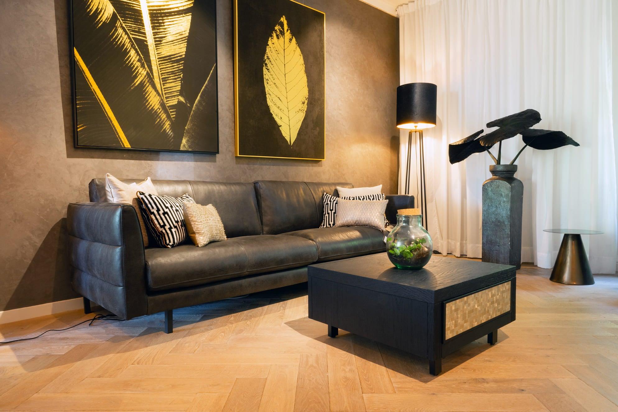 "Stijlvolle zwarte lederen bank met sierkussens in een eigentijds woonkamerinterieur met grote kunstwerken aan de muur, designer vloerlamp, houten vloer en decoratieve elementen."