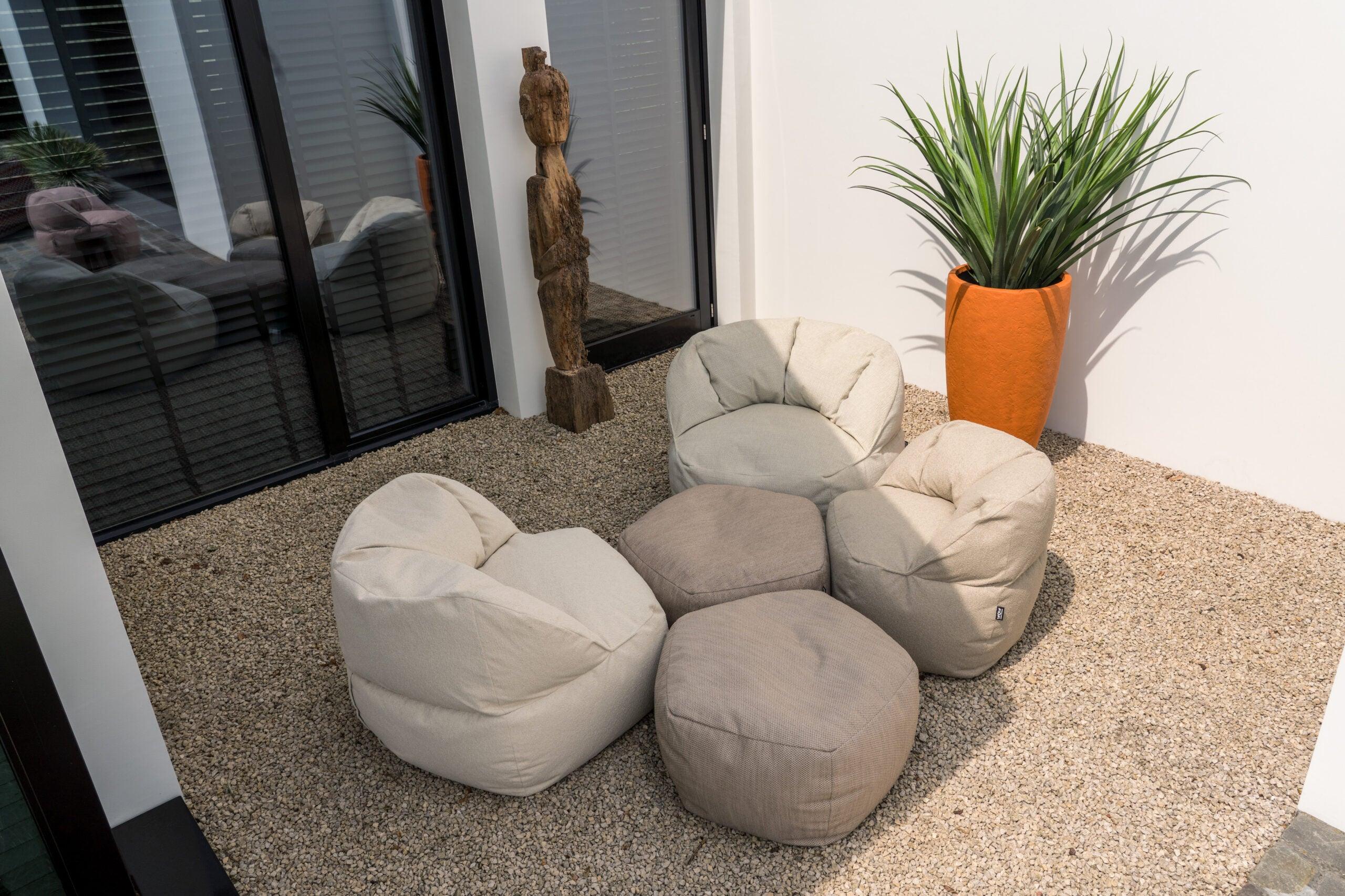 "Moderne terrastuin met comfortabele zitzakken in neutrale kleur, houten sculptuur en grote groene plant in oranje plantenbak naast schuifpui."