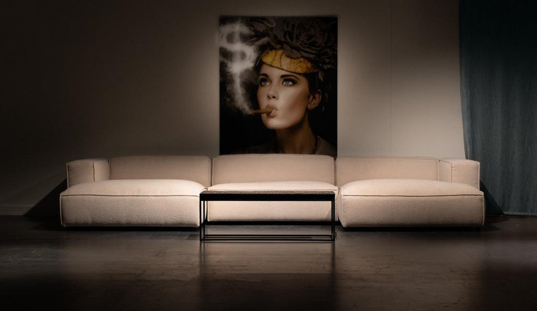 "Stijlvolle beige modulaire hoekbank in een minimalistische woonkamer met artistiek portret van een vrouw met sigaar en hoofdtooi op de achtergrond, verlicht door sfeervolle spots, inclusief een strakke glazen salontafel met metalen frame"