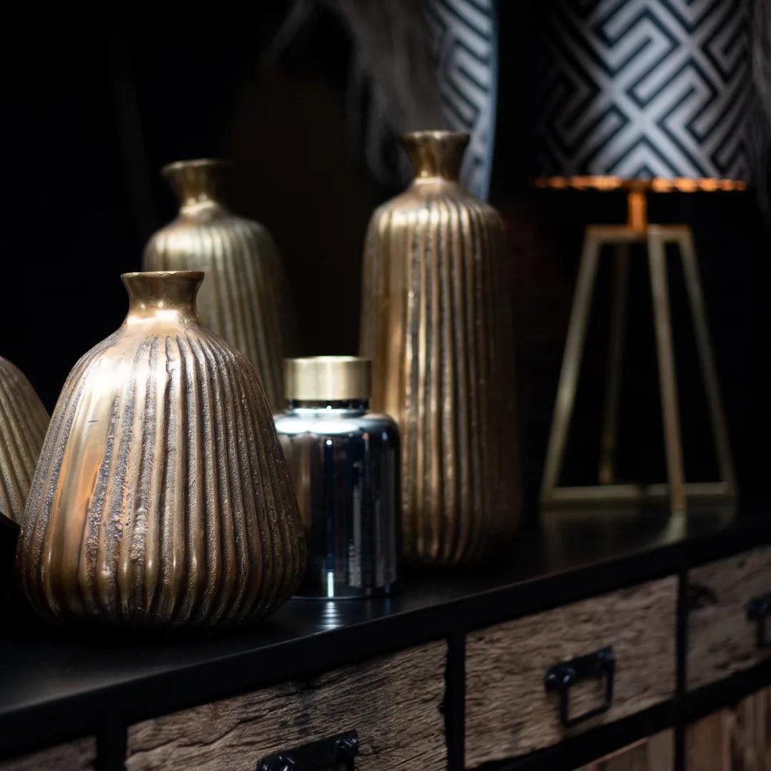 "Gouden decoratieve vazen op een houten console met industriële uitstraling, accentverlichting, en moderne geometrische accessoires op de achtergrond in een sfeervolle interieursetting."