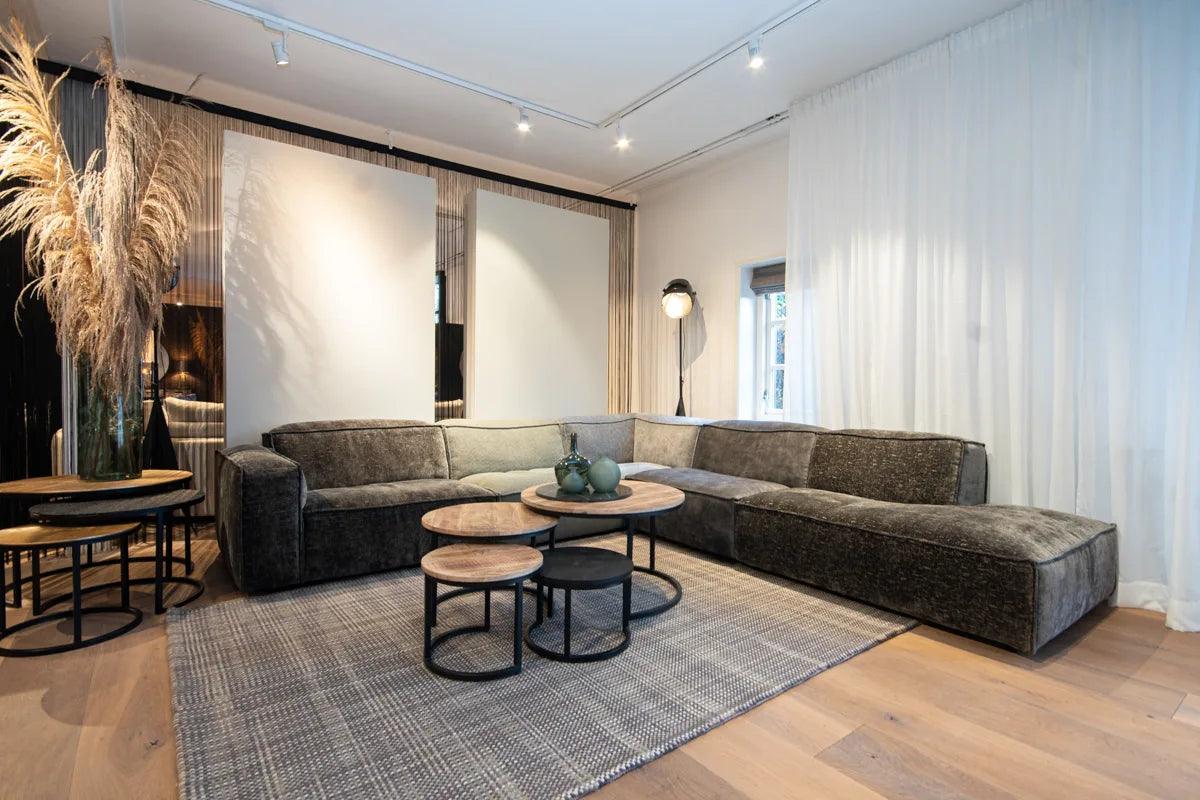 "Interior ontwerp van een stijlvolle woonkamer met een elegante fluwelen hoekbank, ronde houten salontafels, hoge vloerlamp, en decoratieve pampasgras in vaas op een geweven vloerkleed, met natuurlijk licht en strakke witte gordijnen."