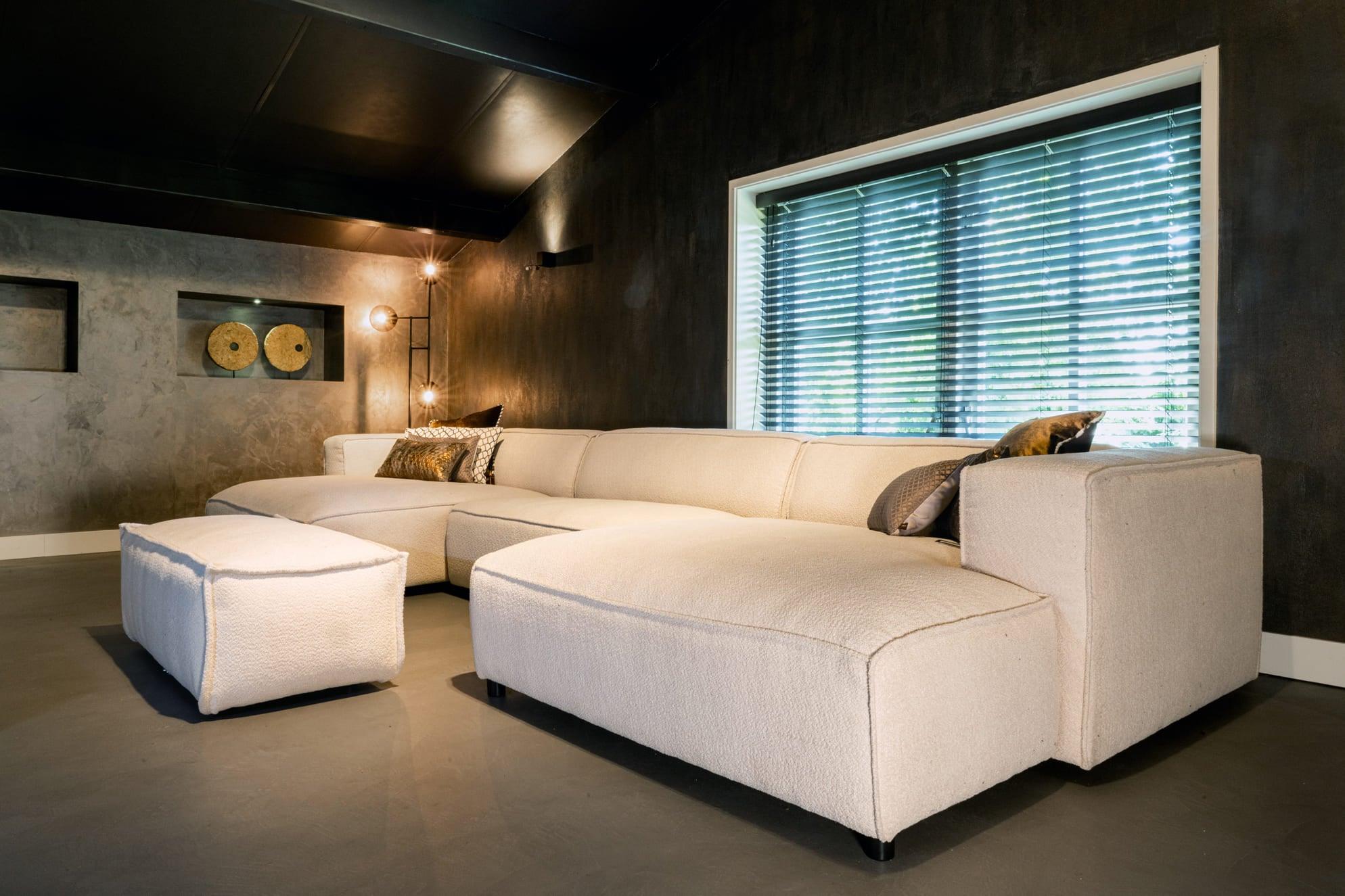 "Design verlichting en decoratieve kussens op stijlvolle beige hoekbank in een luxueus interieur met donkere muren en grote raampartij met horizontale jaloezieën."