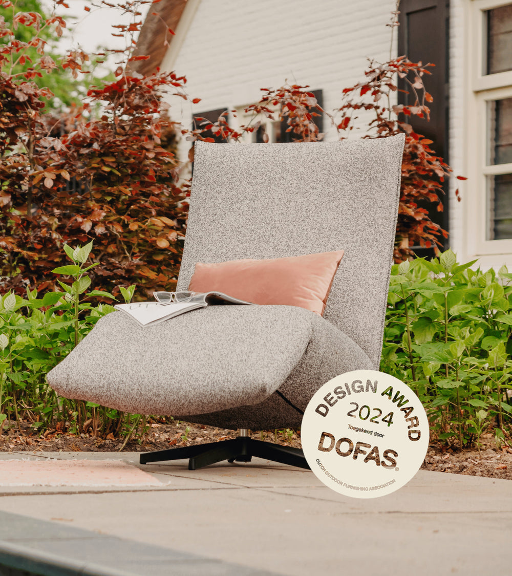 Grijs gemêleerde lounge stoel buiten van HoekbankXXL, met roze kussen, tijdschrift, bril en design award.
