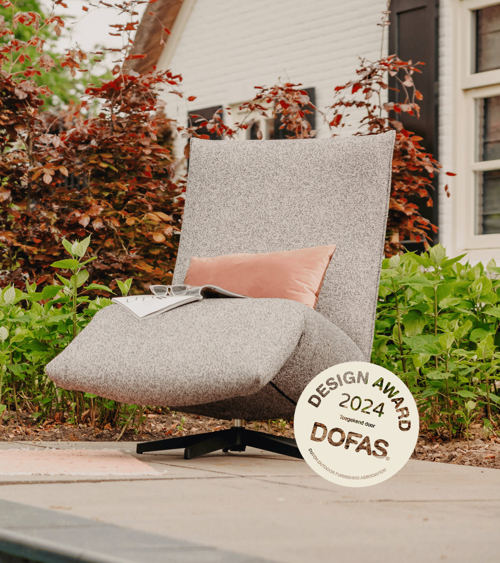 Grijs gemêleerde lounge stoel buiten van HoekbankXXL, met roze kussen, tijdschrift, bril en design award.
