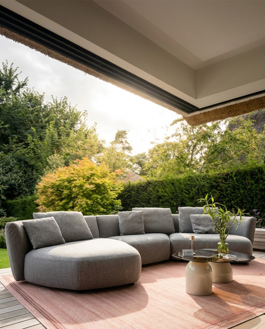 Grijze loungebank tuin Atoll van HoekbankXXL, met zachte kussens op een roze tapijt in een groene omgeving.