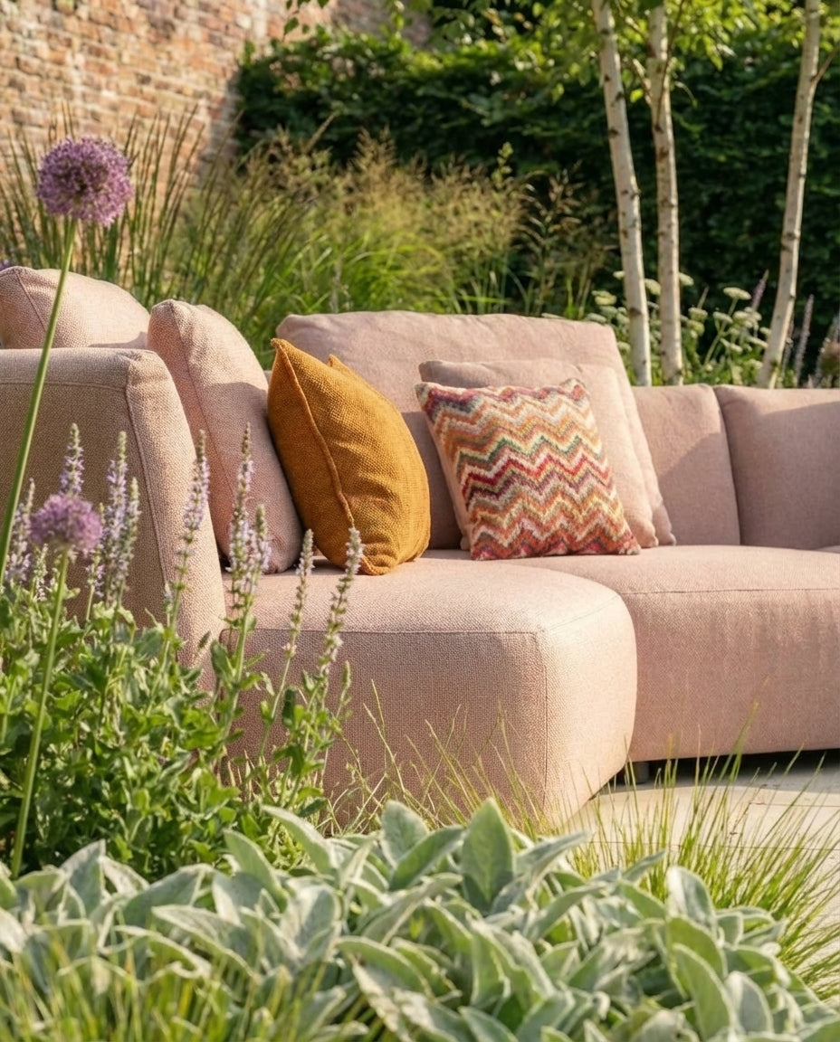 Roze loungebank tuin Atoll van HoekbankXXL, met sierkussens in geel en zigzagpatroon, in een weelderige tuinomgeving.
