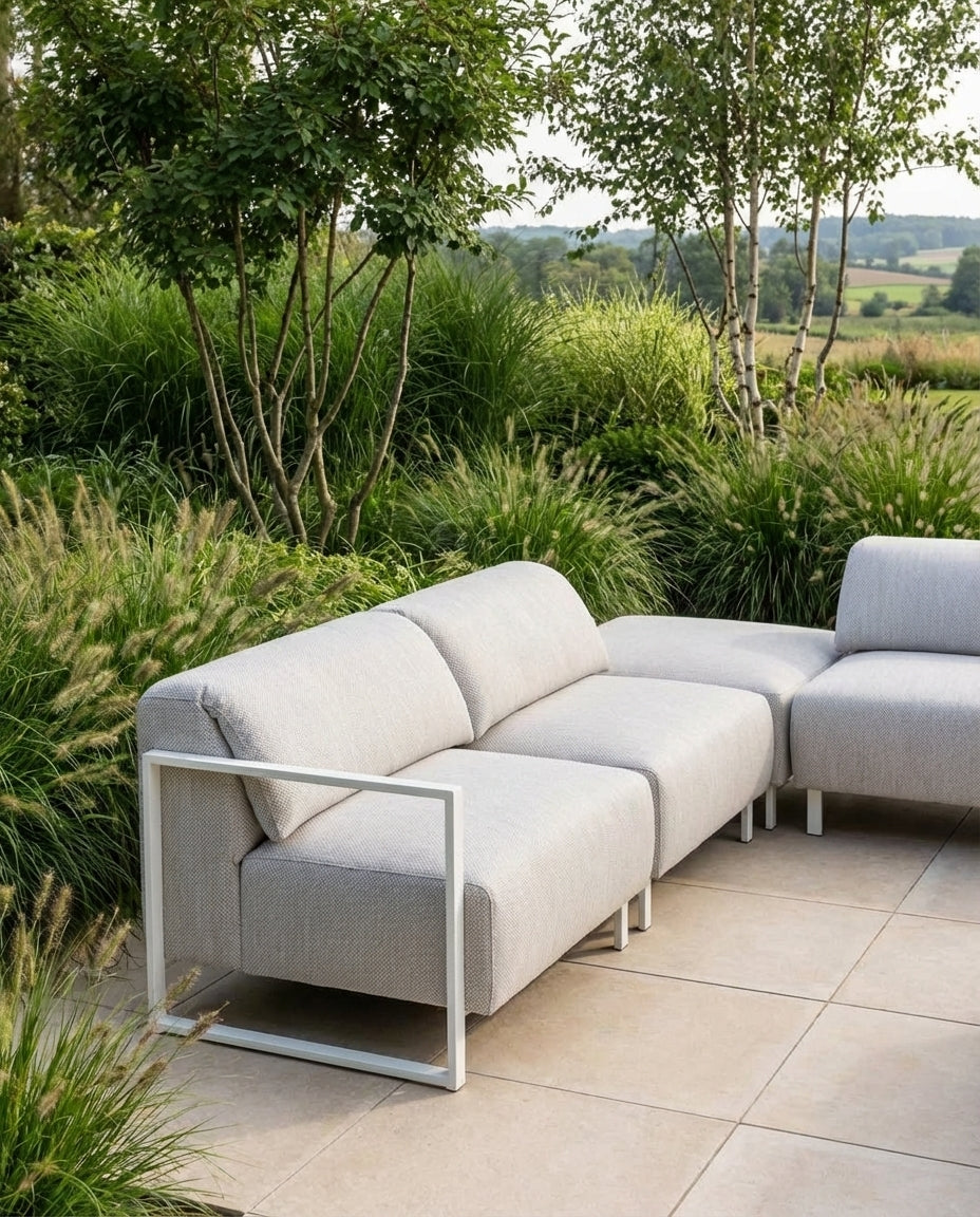 Grijze loungebank tuin Bali van HoekbankXXL, met wit metalen frame, geplaatst op een stenen terras in een groene tuin.
