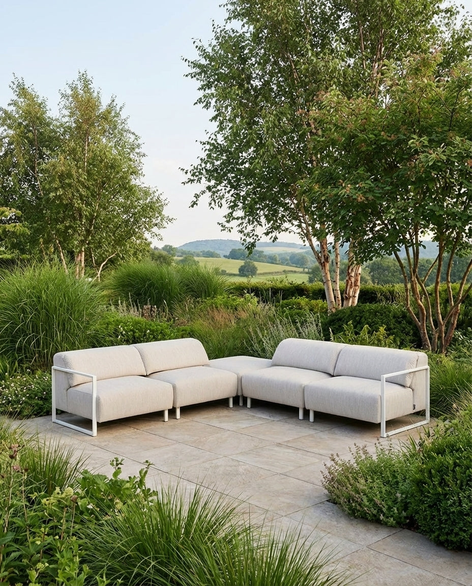 Loungebank Tuin Bali van HoekbankXXL, met een witte frame en lichtgekleurde bekleding, op een stenen terras in een groene tuin.
