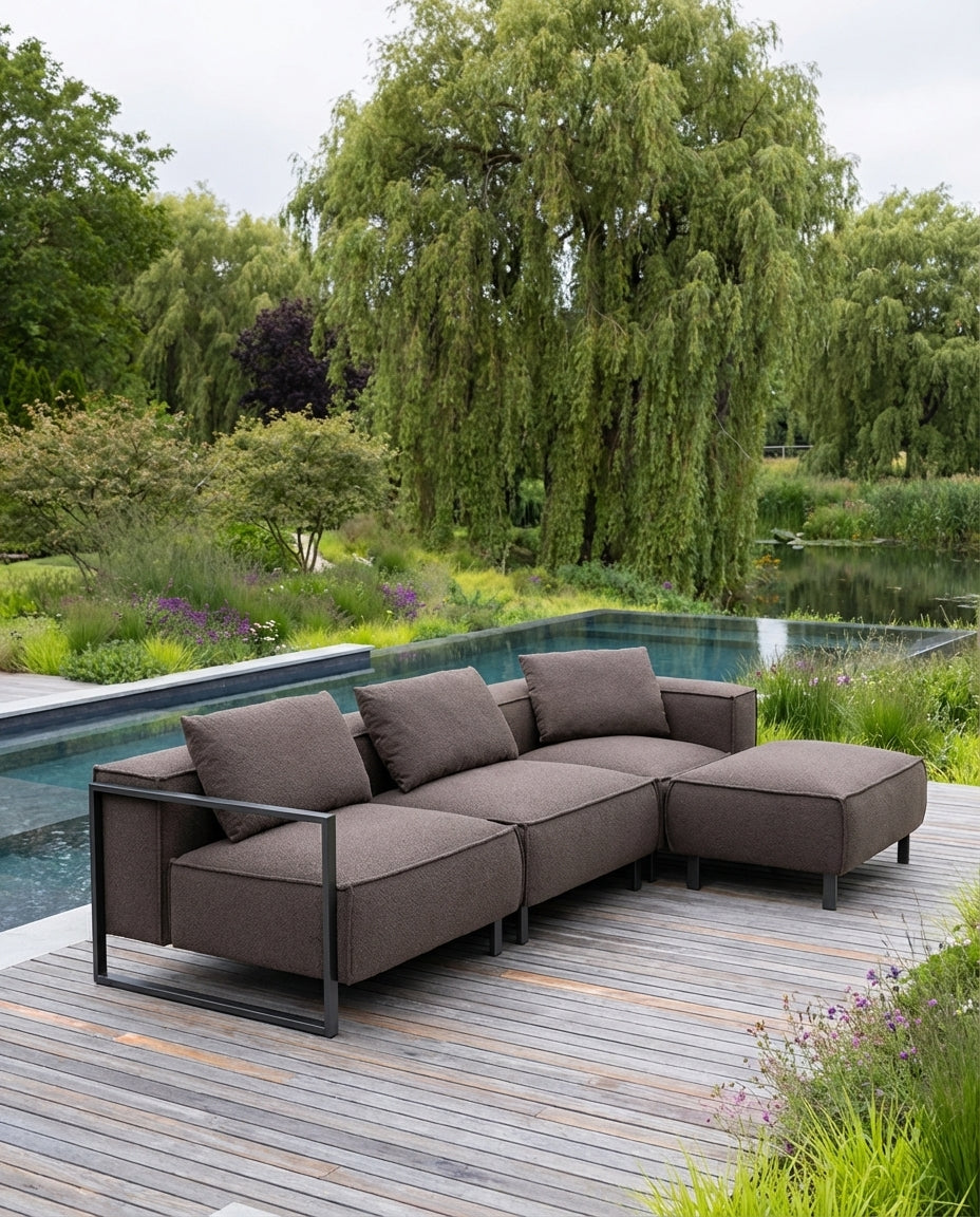 Bruine loungebank tuin van HoekbankXXL, met strakke metalen frame en comfortabele kussens, op houten terras bij water.

