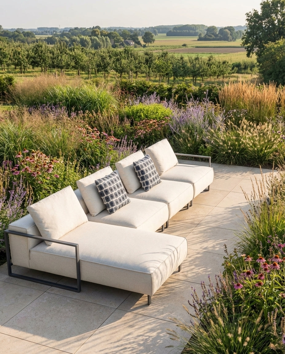 Crèmekleurige loungebank tuin - Mauritius van HoekbankXXL met sierkussens op een betegelde patio in een weelderige tuin.
