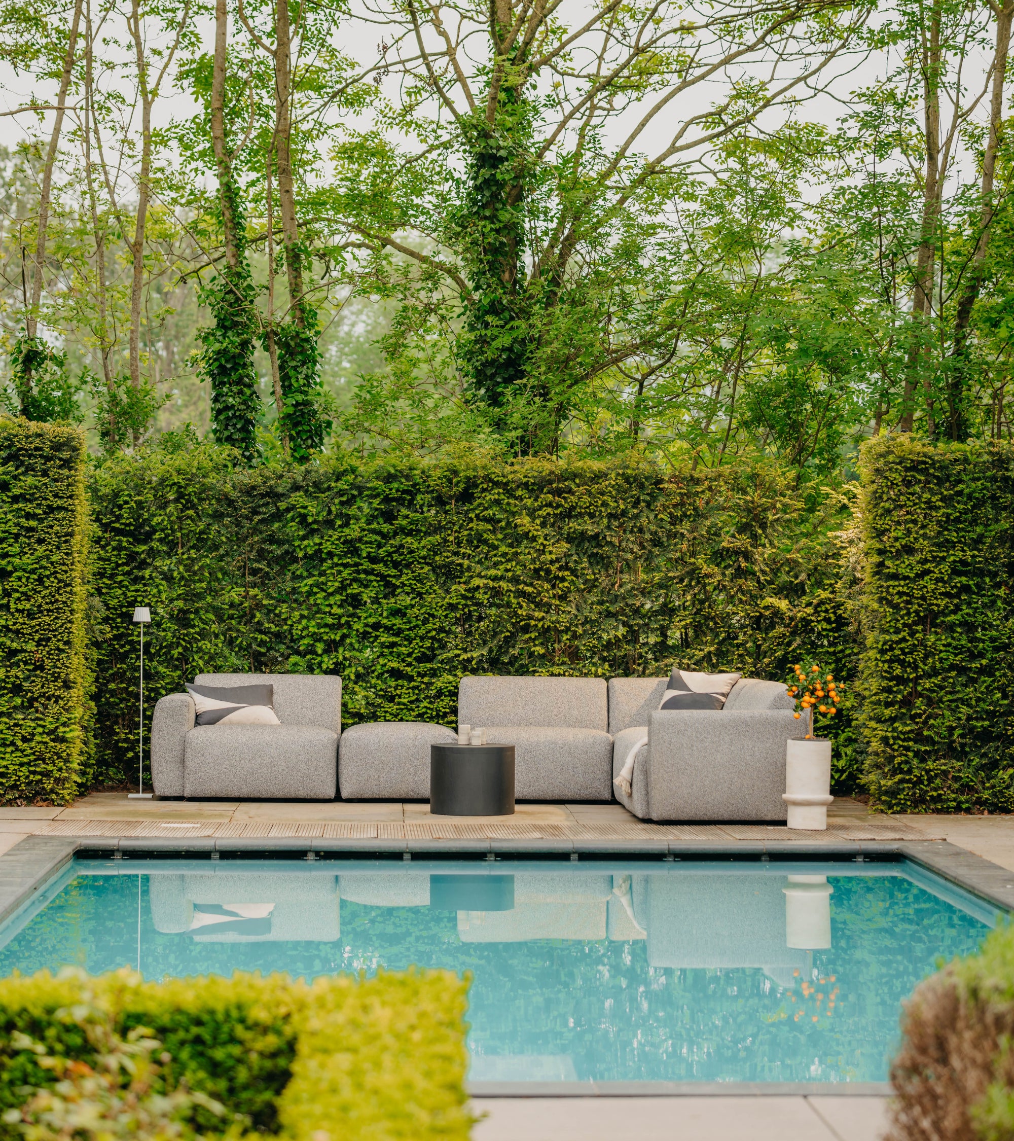 Grijze loungebank tuin van HoekbankXXL, met kussens en bijzettafel bij een zwembad in een groene tuin.
