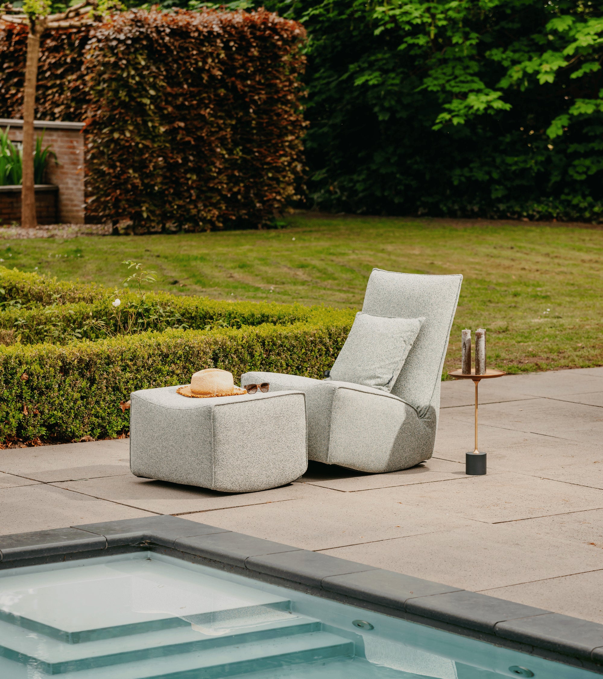 Grijze lounge fauteuil van HoekbankXXL met kussen en bijpassende hocker, compleet met hoed en zonnebril op een terras bij een zwembad.
