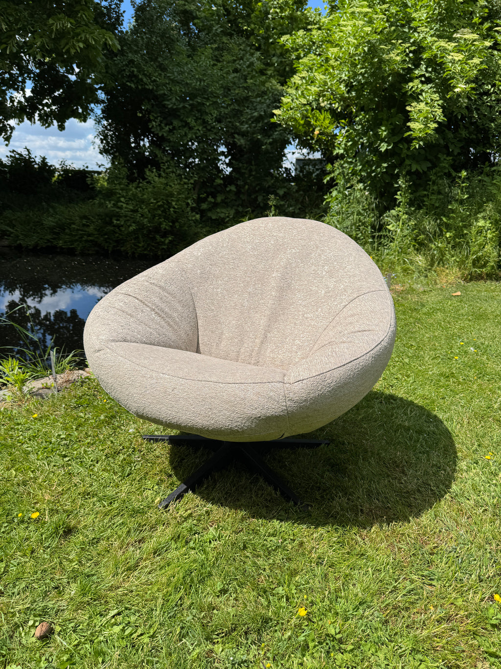 Beige outdoor fauteuil Bora Bora van HoekbankXXL, met een afgeronde vorm en een zwart onderstel, staat op een gazon in de tuin.
