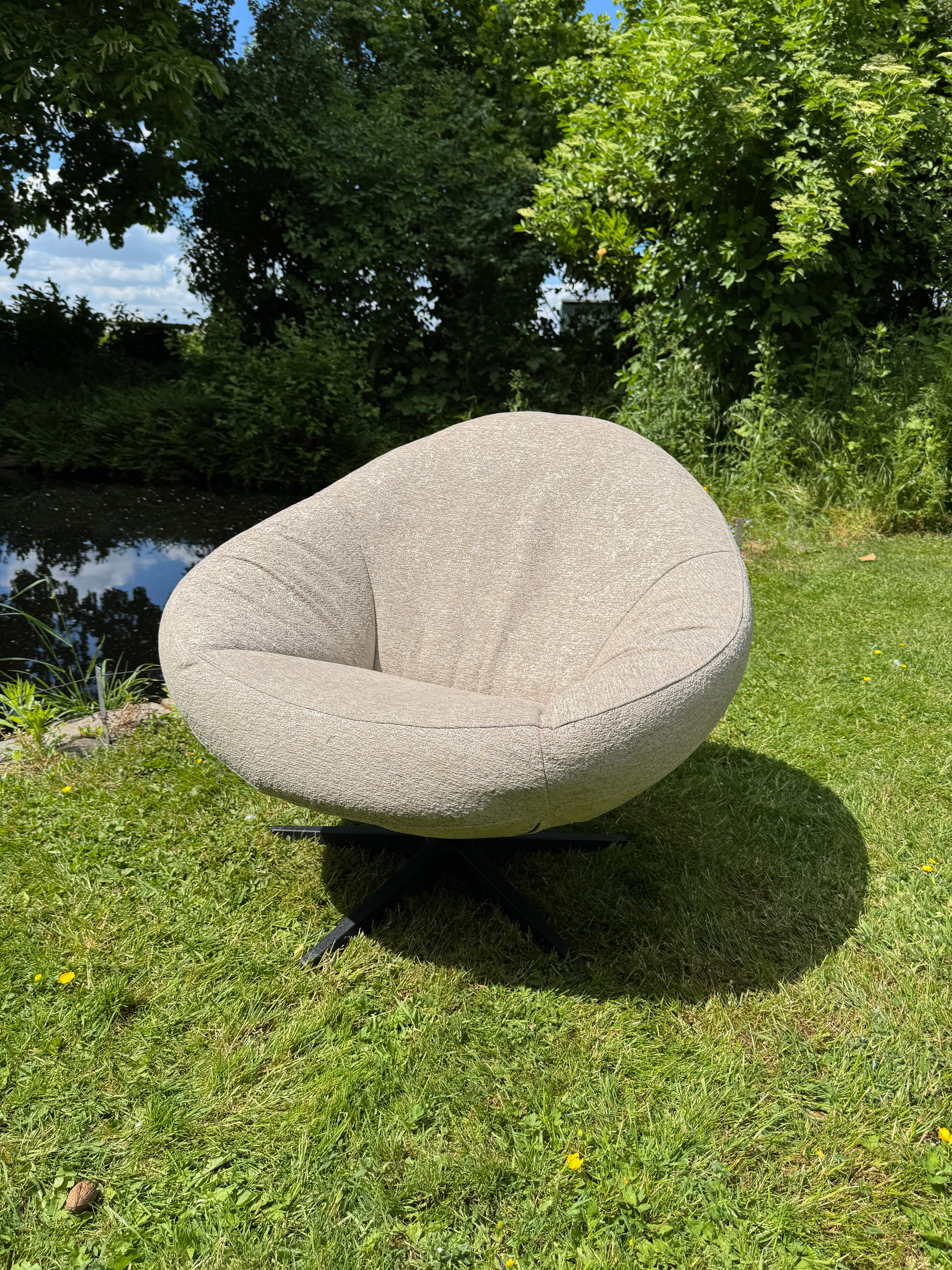 Beige outdoor fauteuil Bora Bora van HoekbankXXL, met een afgeronde vorm en een zwart onderstel, staat op een gazon in de tuin.

