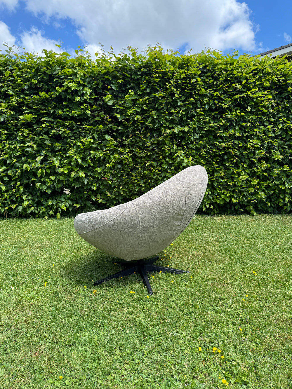 Grijze outdoor fauteuil Bora Bora van HoekbankXXL, met een zwart onderstel, geplaatst op een gazon met klaver en paardenbloemen.
