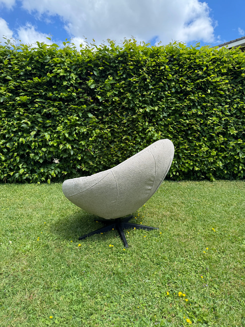 Grijze outdoor fauteuil Bora Bora van HoekbankXXL, met een zwart onderstel, geplaatst op een gazon met klaver en paardenbloemen.
