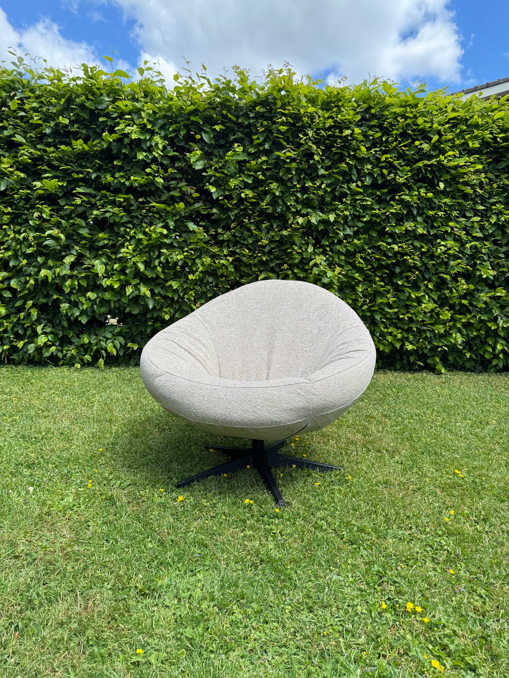 Grijze fauteuil Bora Bora van HoekbankXXL, met een ronde zitting en zwart onderstel, staand op groen gras met bloemen.
