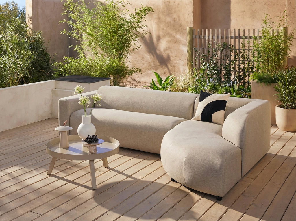 Beige Penna outdoor loungebank van HoekbankXXL, met een zwart-wit kussen en een bijpassende ronde tafel met accessoires.
