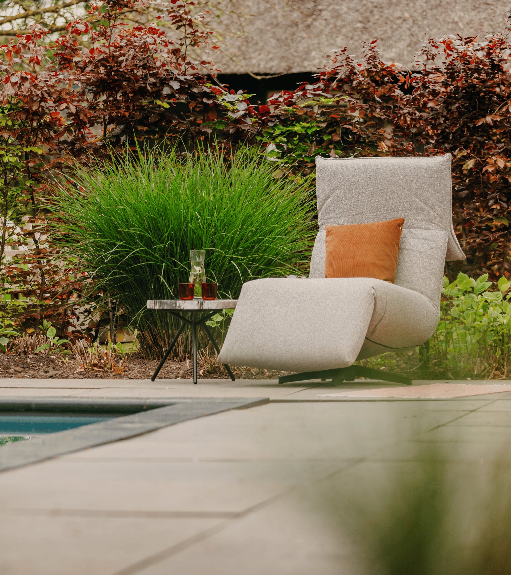 Grijze lounge stoel buiten van HoekbankXXL, met een bruin kussen, naast een klein marmeren tafeltje.
