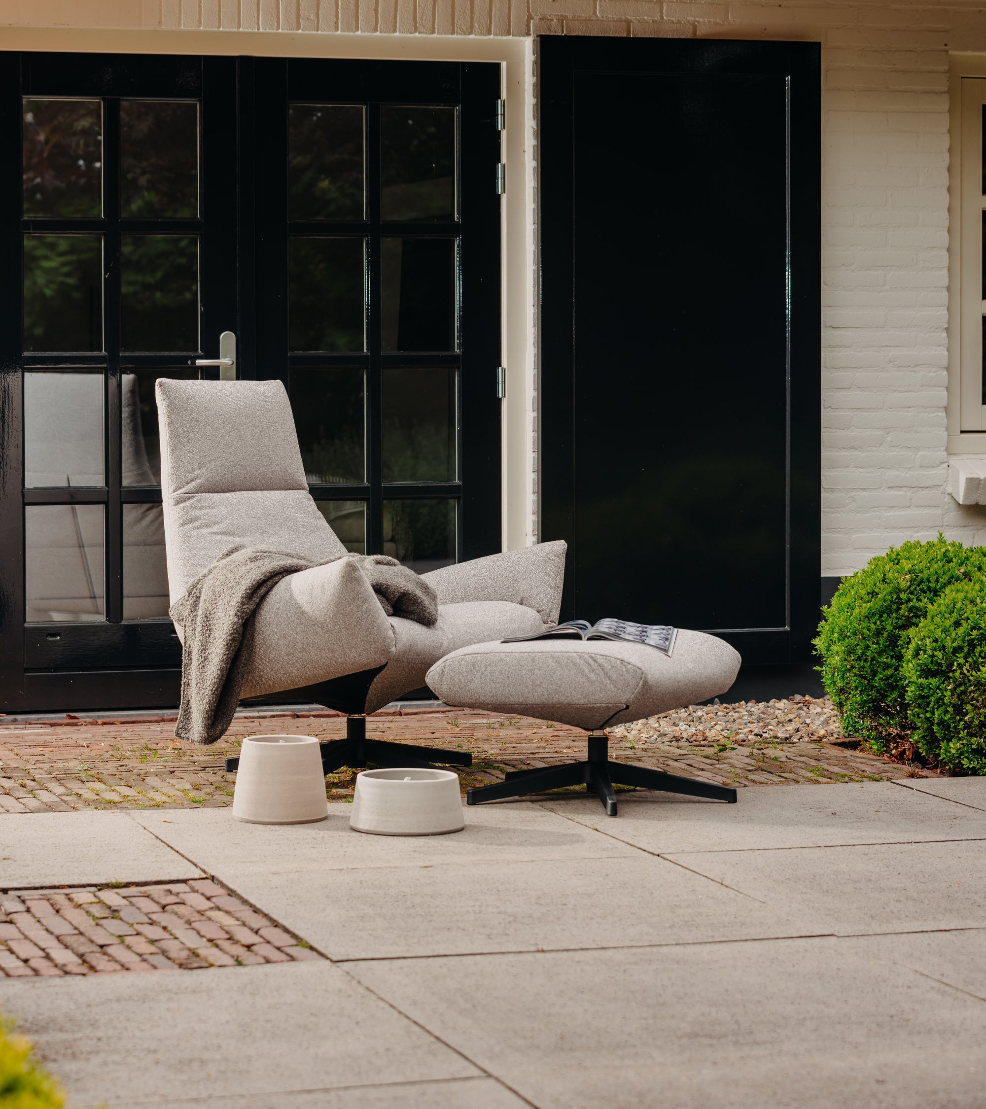 Sicilië Lounge Fauteuil van HoekbankXXL met een lichtgrijze stof, vergezeld van een bijpassende voetenbank en twee decoratieve accessoires.

