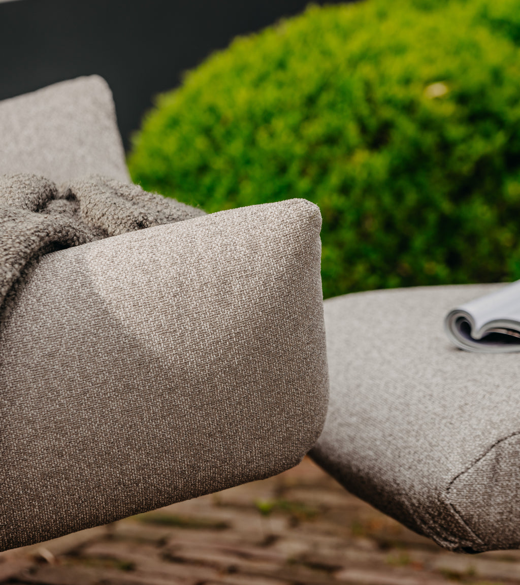 Grijze lounge fauteuil van HoekbankXXL met grof geweven textuur, een zachte deken en tijdschrift erop, in de tuin.
