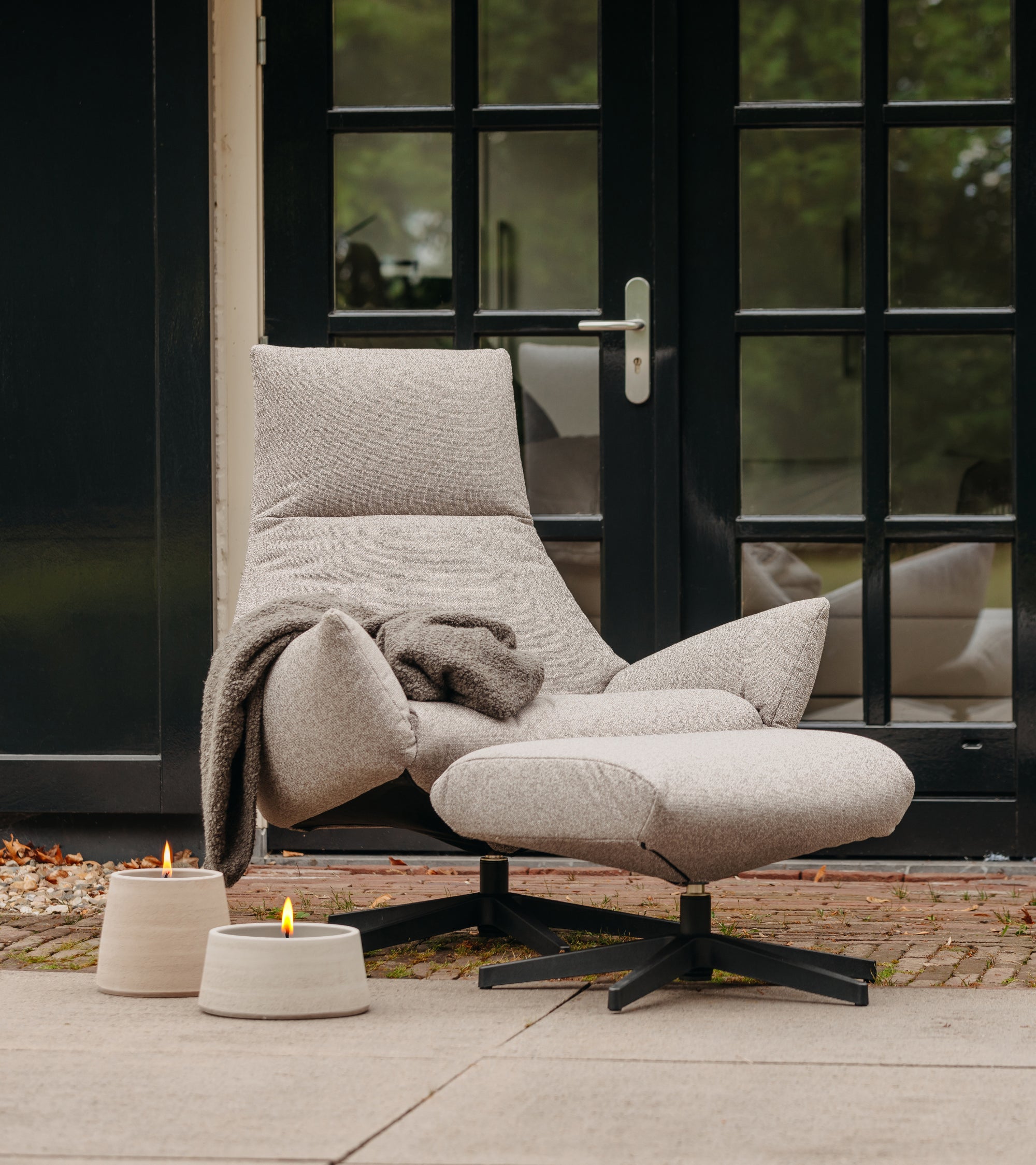 Sicily lounge fauteuil van HoekbankXXL, met voetbank en een bruin dekentje, naast twee kaarsen op een terras.
