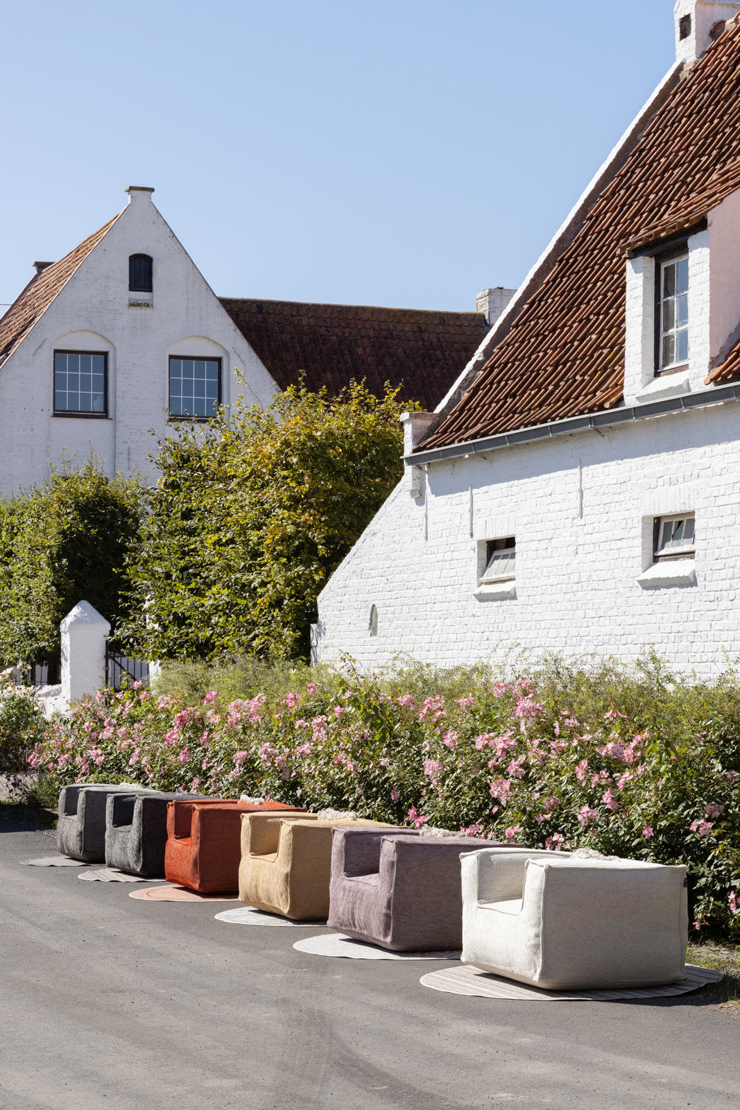 Silky clubchair loungestoel buiten van HoekbankXXL, in diverse kleuren opgesteld voor witte gebouwen en bloeiende struiken.
