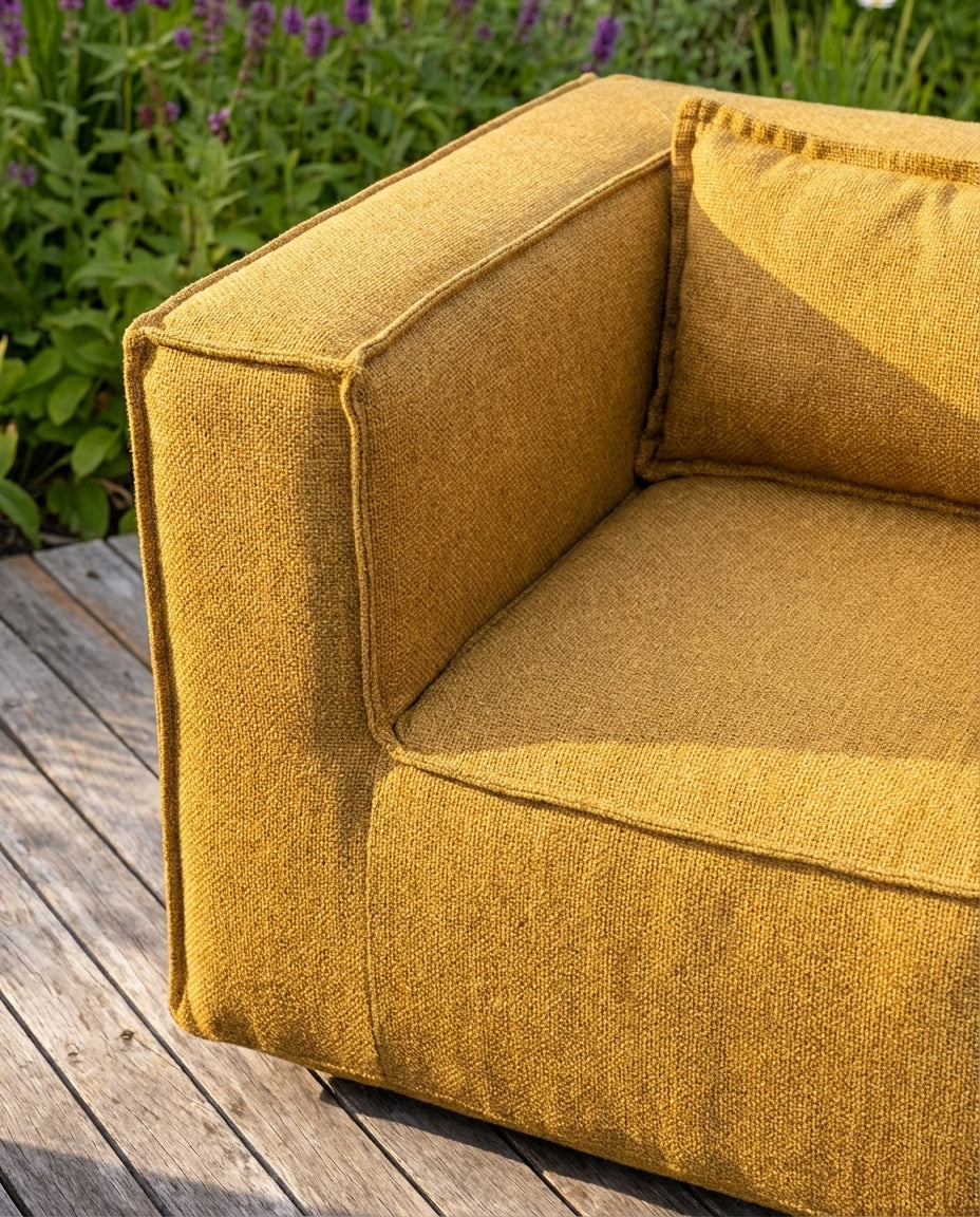 Gele lounge stoel buiten, de Silky Clubchair van HoekbankXXL, met een grof geweven textuur op een houten terras.
