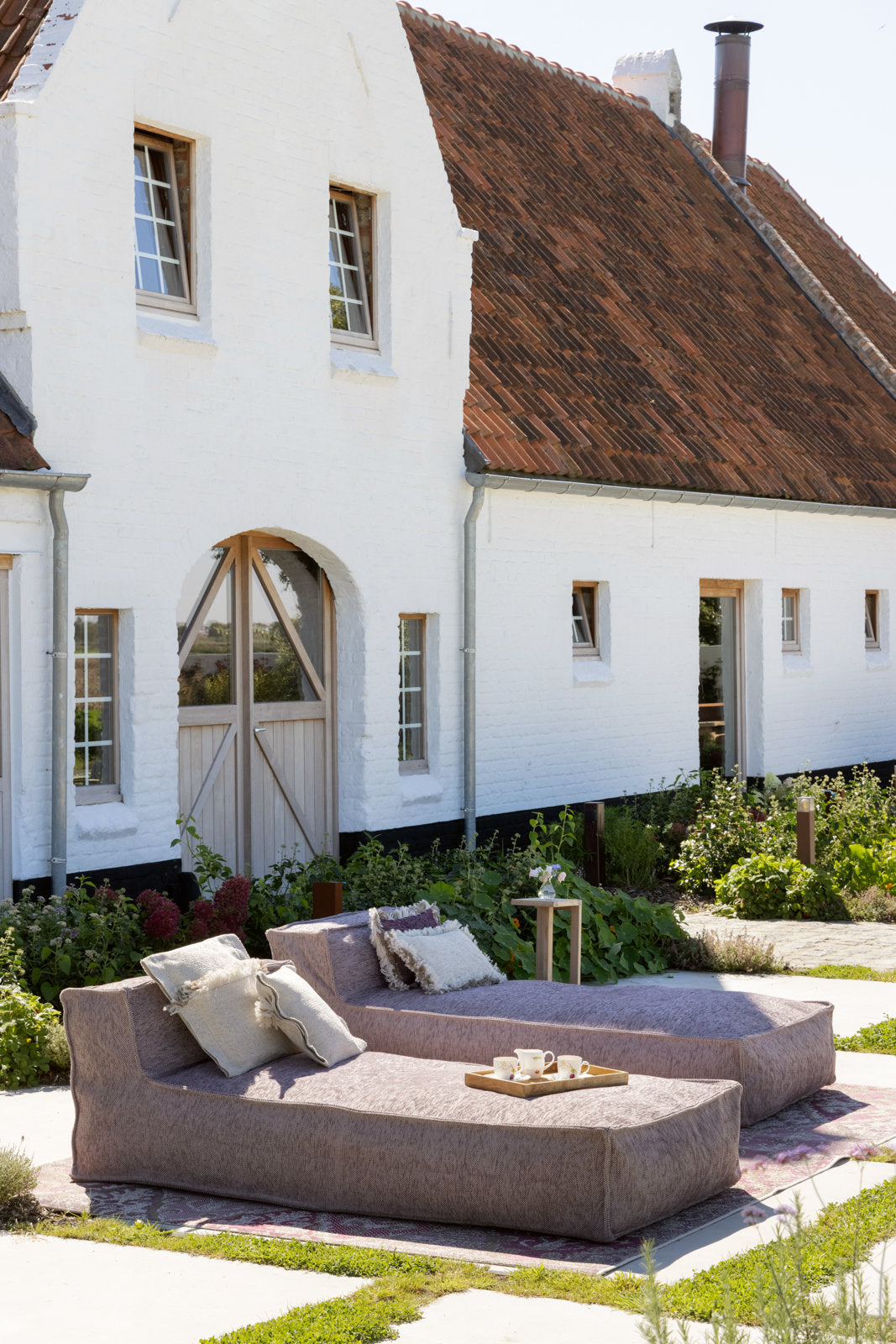 Zijdeachtige longchair loungestoel voor buiten van HoekbankXXL, met zachte kussens en een houten dienblad, geplaatst op een decoratief tapijt in een tuin.
