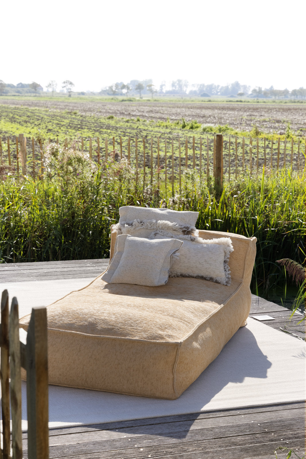 Zandkleurige silky longchair loungestoel buiten van HoekbankXXL, met zachte kussens, op een houten terras in een landelijke omgeving.
